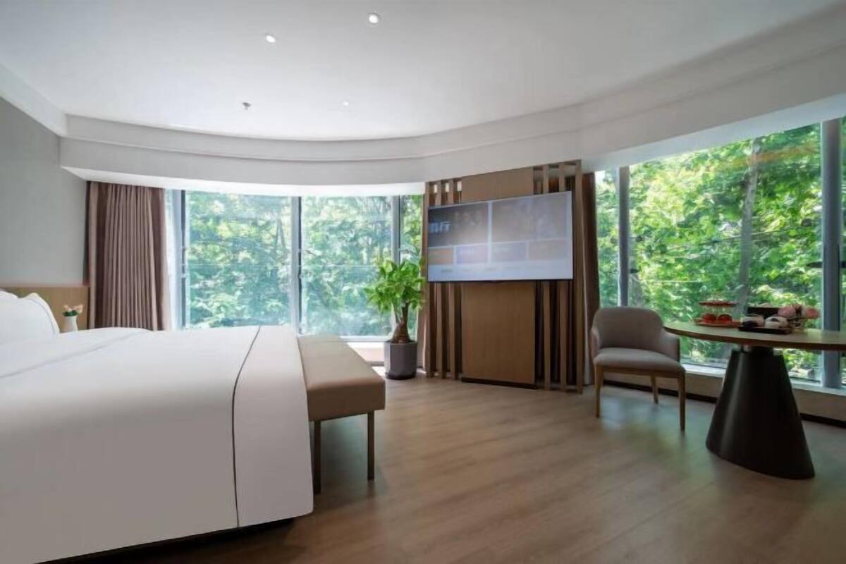 Ein modernes Hotelzimmer mit einem großen Bett, einem Fernseher, einem Esstisch und Blick auf Grün durch große Fenster.