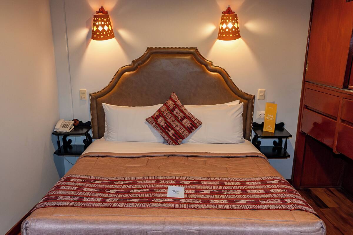 A hotel room with a large bed, two bedside tables, a bedside lamp, a telephone, a bedside box, and a bench.