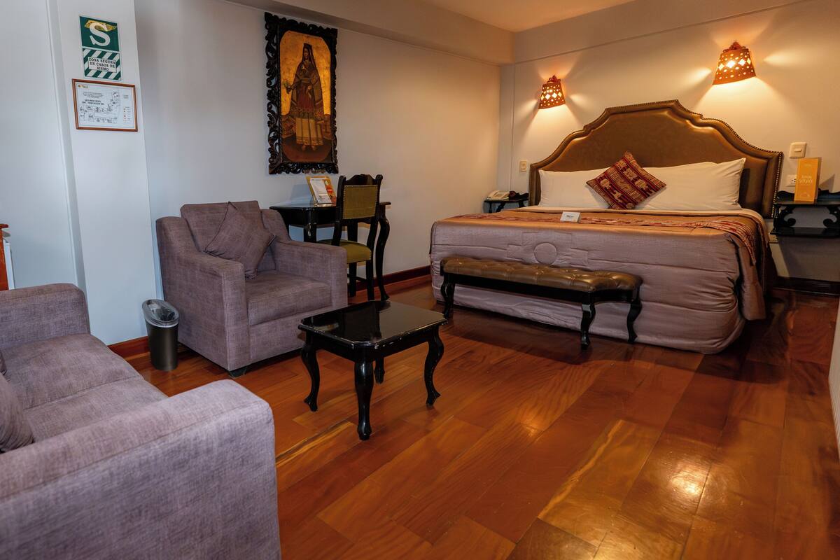 A hotel room with a bed, two armchairs, a small table, a wooden floor, and wall-mounted lights.