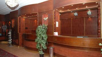 A reception area with a wooden reception desk, a potted plant, a trash can, and a chandelier.