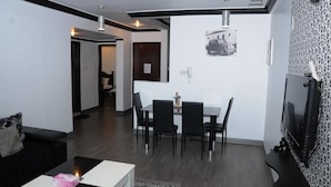 Una sala de estar moderna con un sofá negro, una mesa de centro y un televisor. Hay un comedor con una mesa y sillas. La habitación tiene un papel tapiz estampado y una lámpara en el techo.