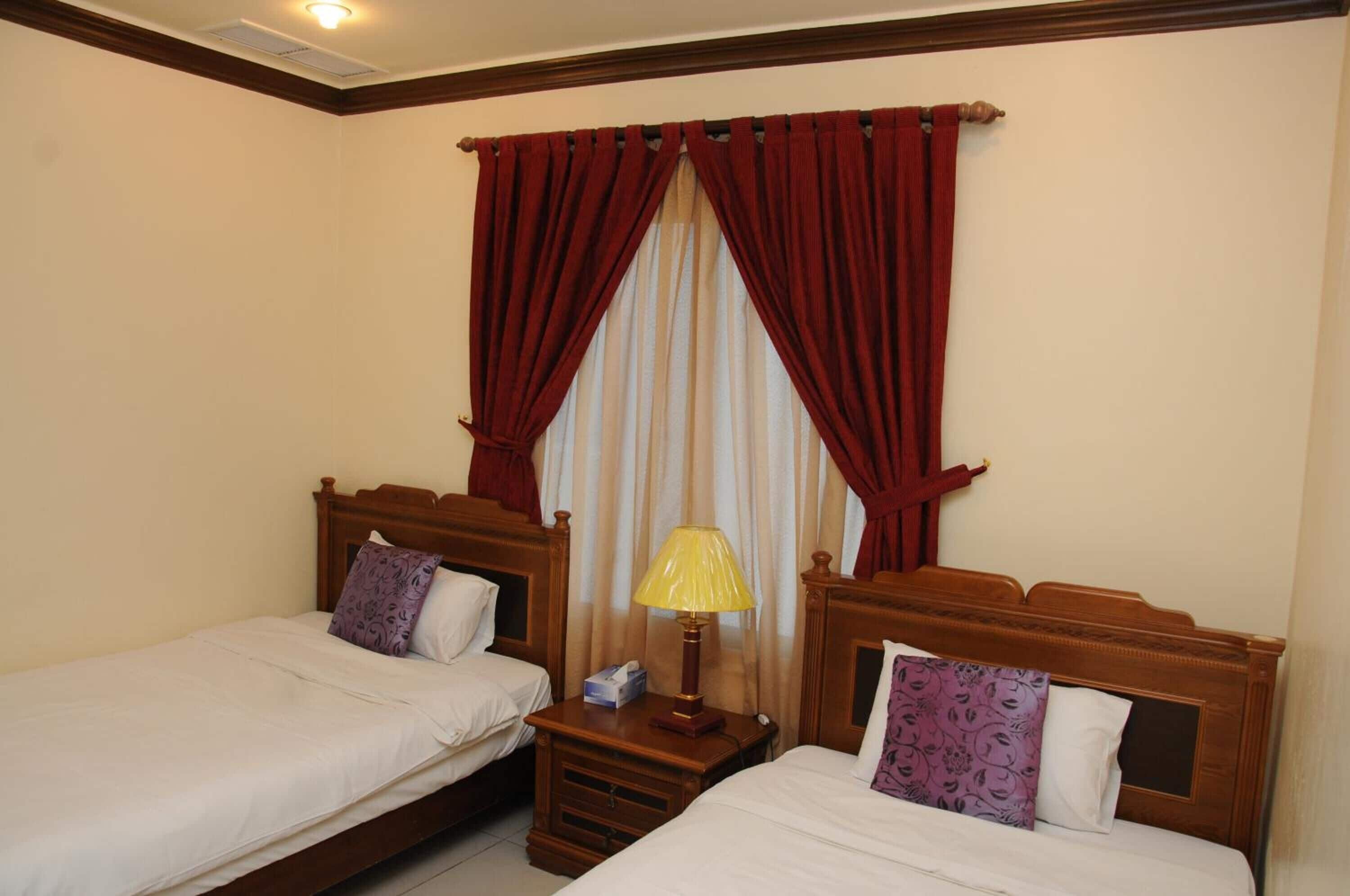 Une chambre d’hôtel avec deux lits simples, une tête de lit en bois, une table de chevet avec une lampe et une fenêtre avec des rideaux rouges.