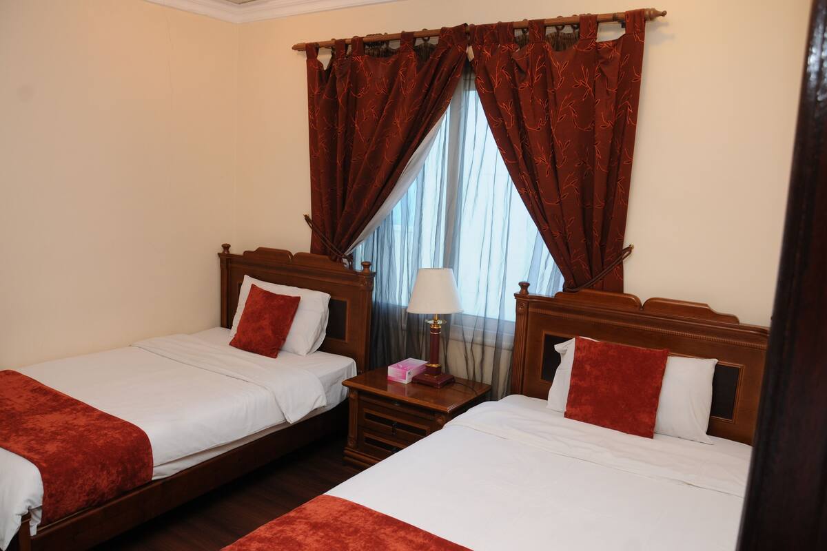 Une chambre d’hôtel avec deux lits simples, chacun doté d’une tête de lit en bois, de draps blancs et d’éléments décoratifs rouges. Il y a une table de chevet avec une lampe et une boîte entre les lits. La chambre est équipée d’une grande fenêtre avec des rideaux rouges.