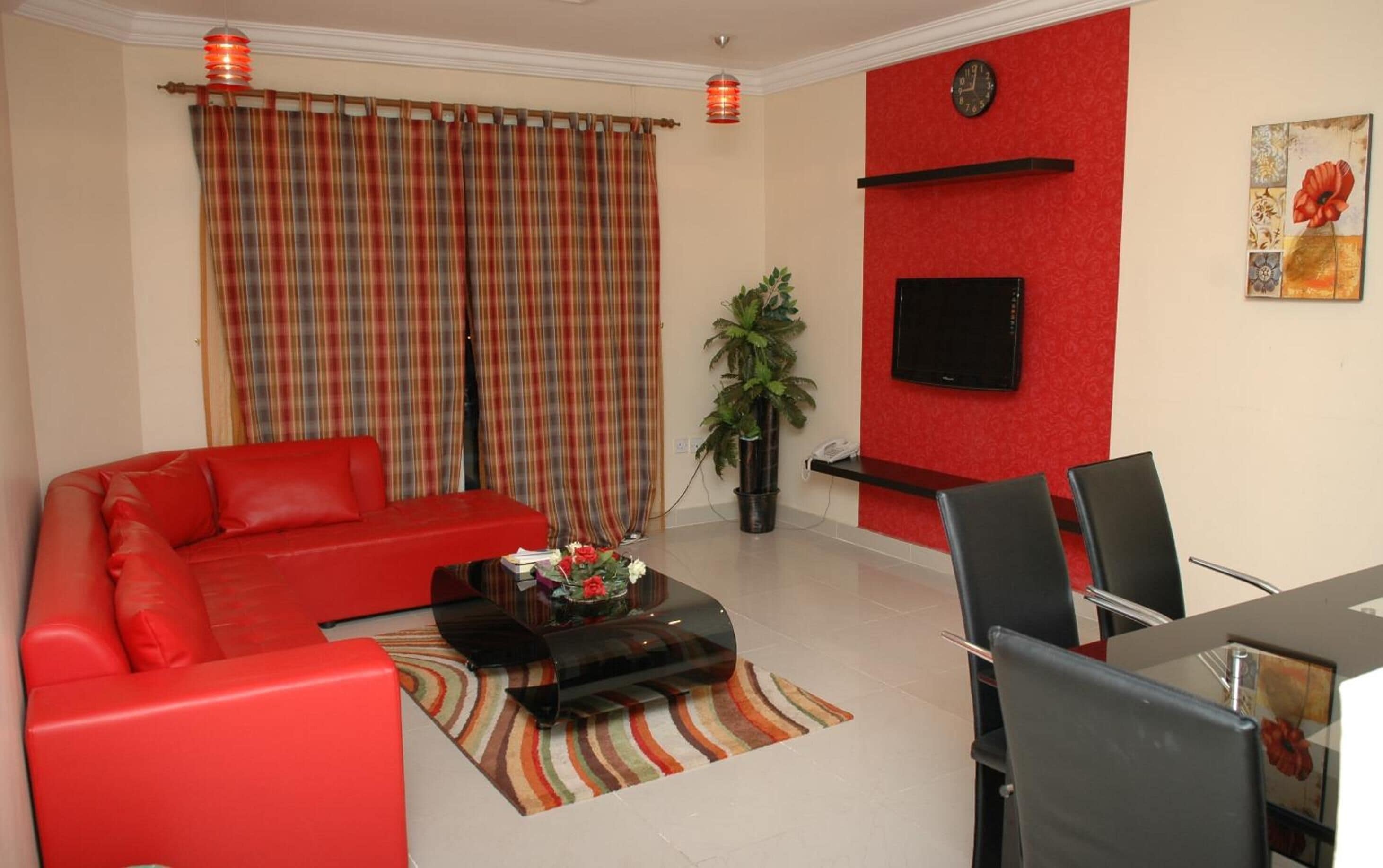 Un salon avec un canapé rouge, une table basse ornée de fleurs, un téléviseur fixé à un mur rouge et un coin repas avec des chaises.