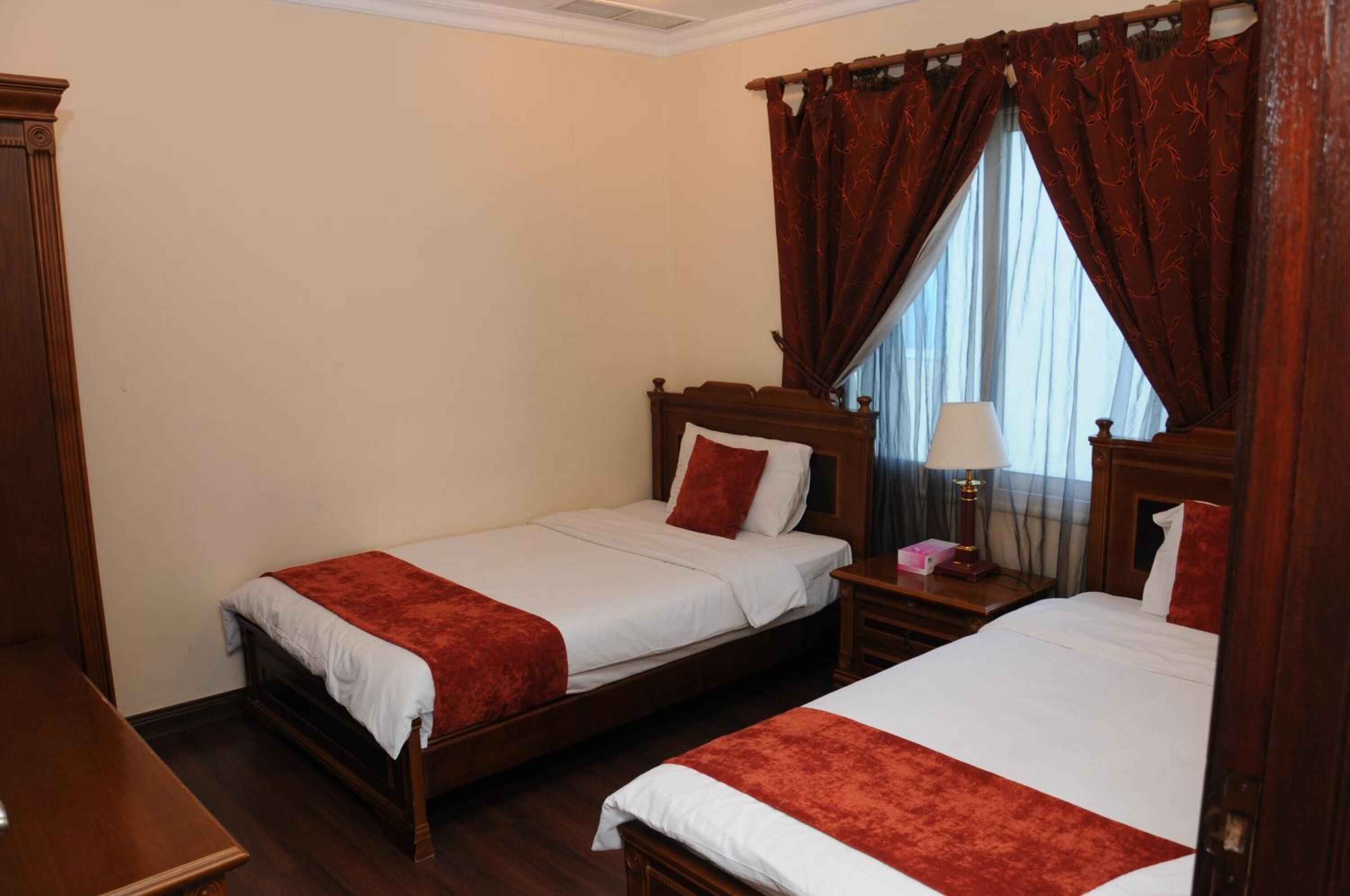 A hotel room with two single beds, wooden headboards, a nightstand with a lamp, and a window with red curtains.