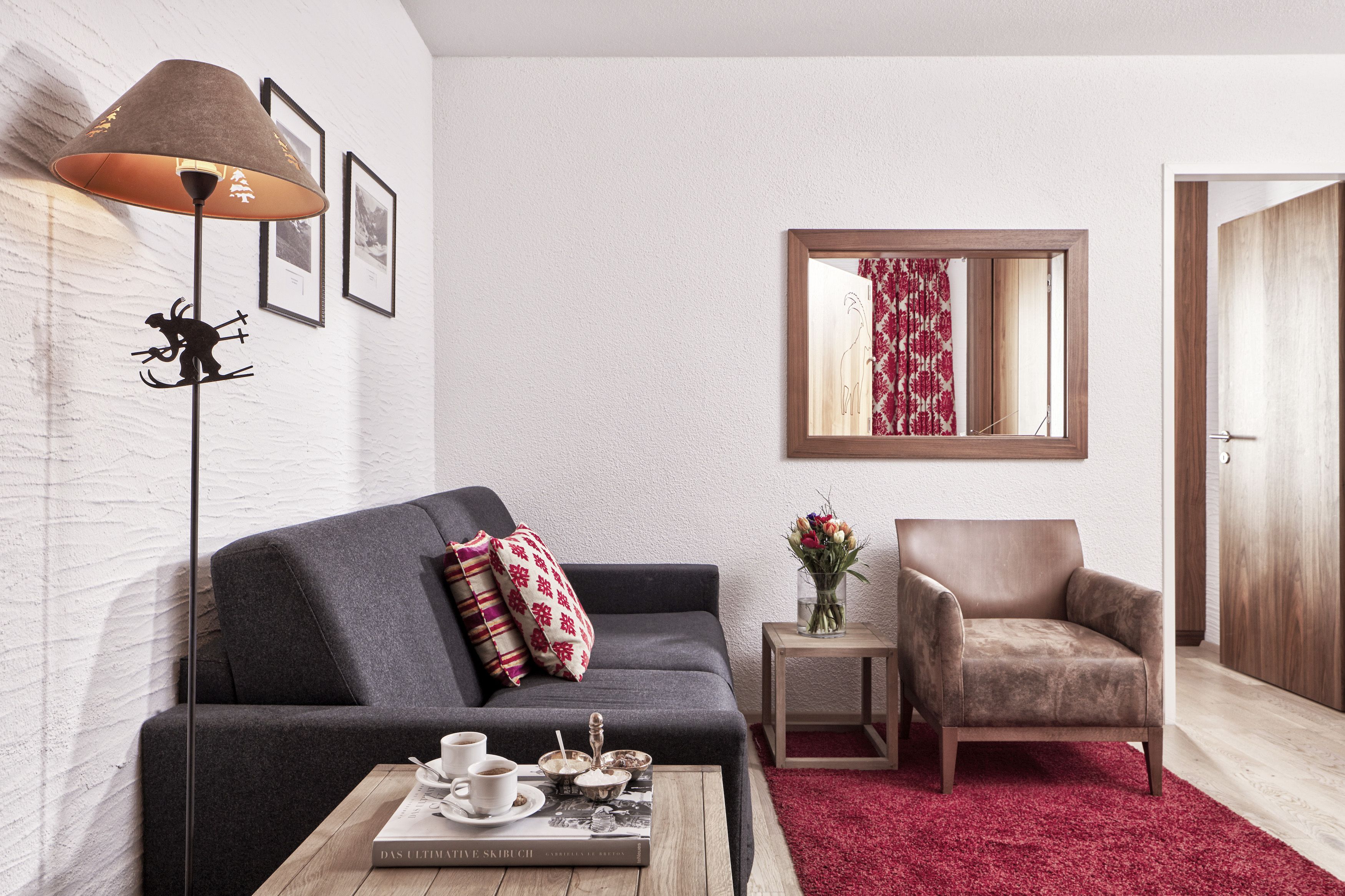 A modern living room with a grey sofa, a small side table with a vase, a red rug, and a wooden floor.