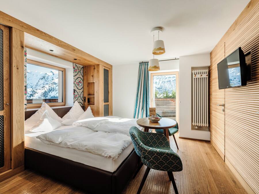 Un dormitorio con cama, una silla, una mesa, un televisor y una ventana con vistas a montañas nevadas.