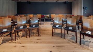Un salón de conferencias con filas de sillas y mesas de madera, una pantalla negra al frente y un piso con diseño.