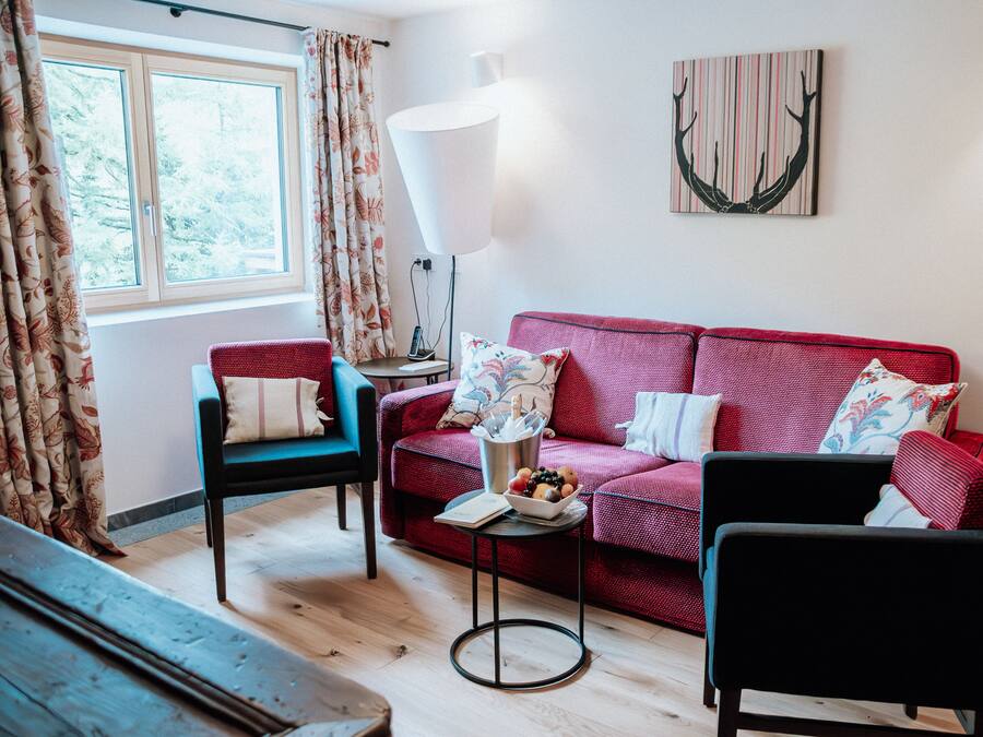 A cozy living room with a red sofa, a blue armchair, a small table with a lamp, and a window with curtains.