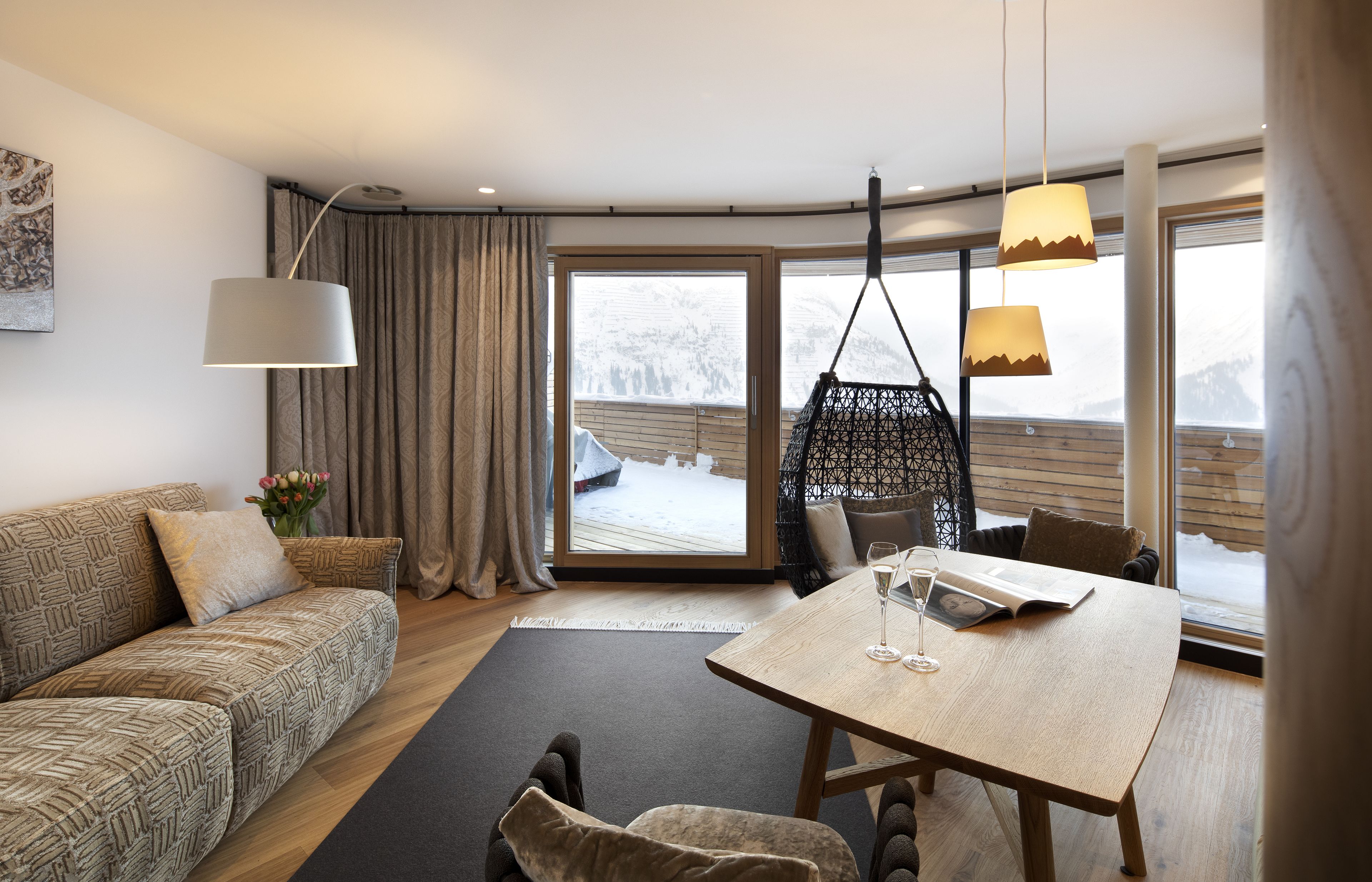 A modern living room with a sofa, a dining table, and a hanging chair. A large window offers a view of a snowy deck outside.