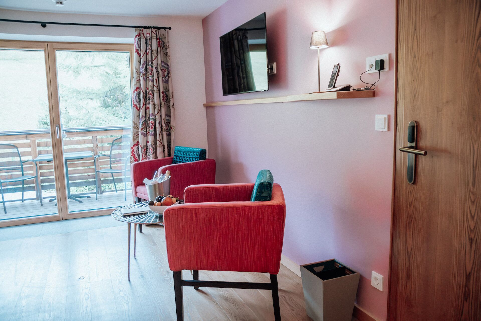 A room with a sliding glass door leading to an outdoor area, a red sofa, a small table, and a mounted TV.