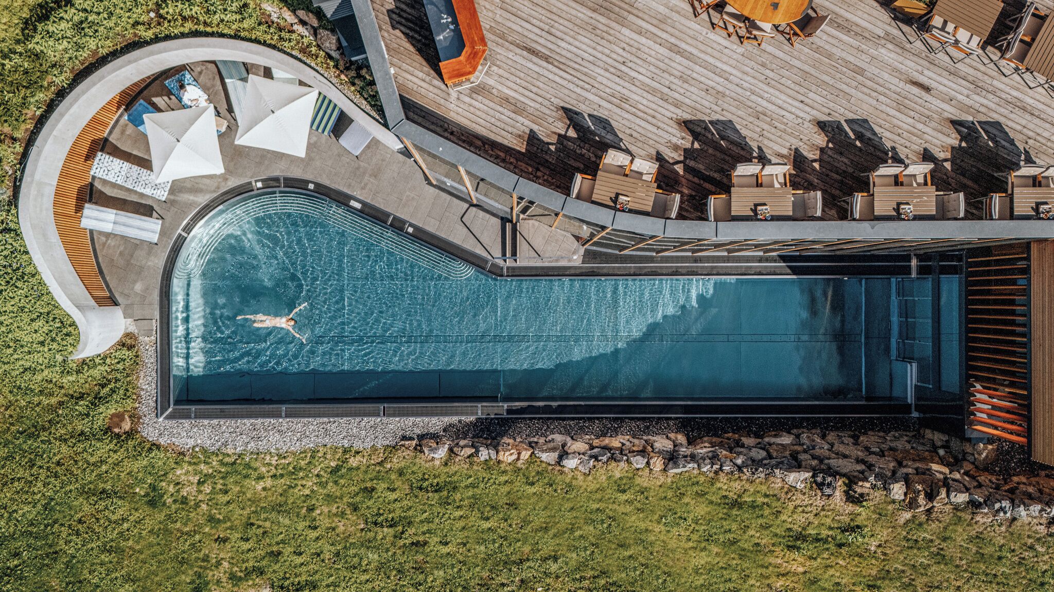 Eine Vogelperspektive auf ein modernes Schwimmbecken mit geschwungener Form, umgeben von Liegestühlen und einer Holzterrasse.