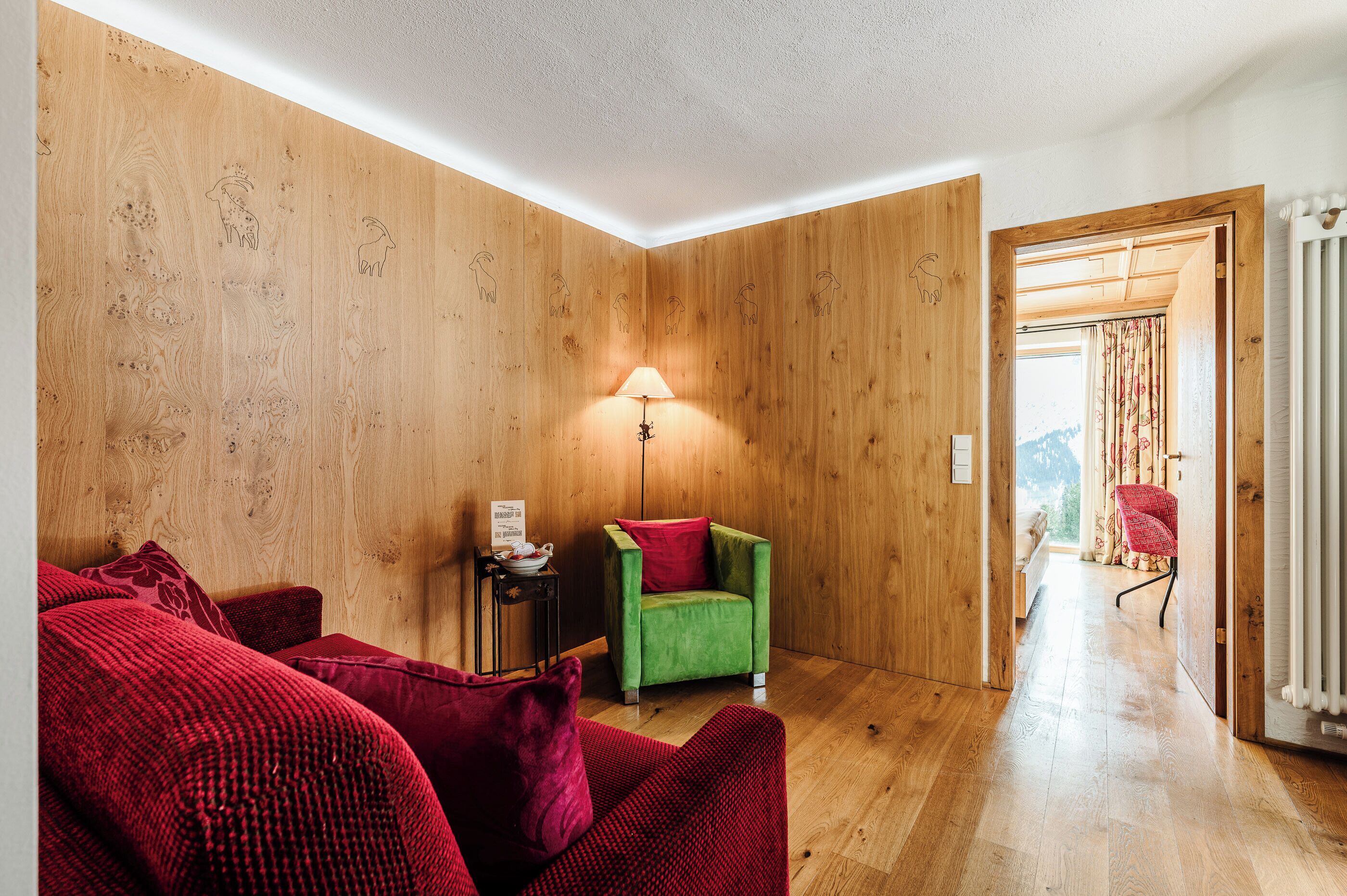 Une pièce aux murs en bois, avec un fauteuil vert, des coussins rouges, une lampe et une vue sur l’extérieur par une porte ouverte.