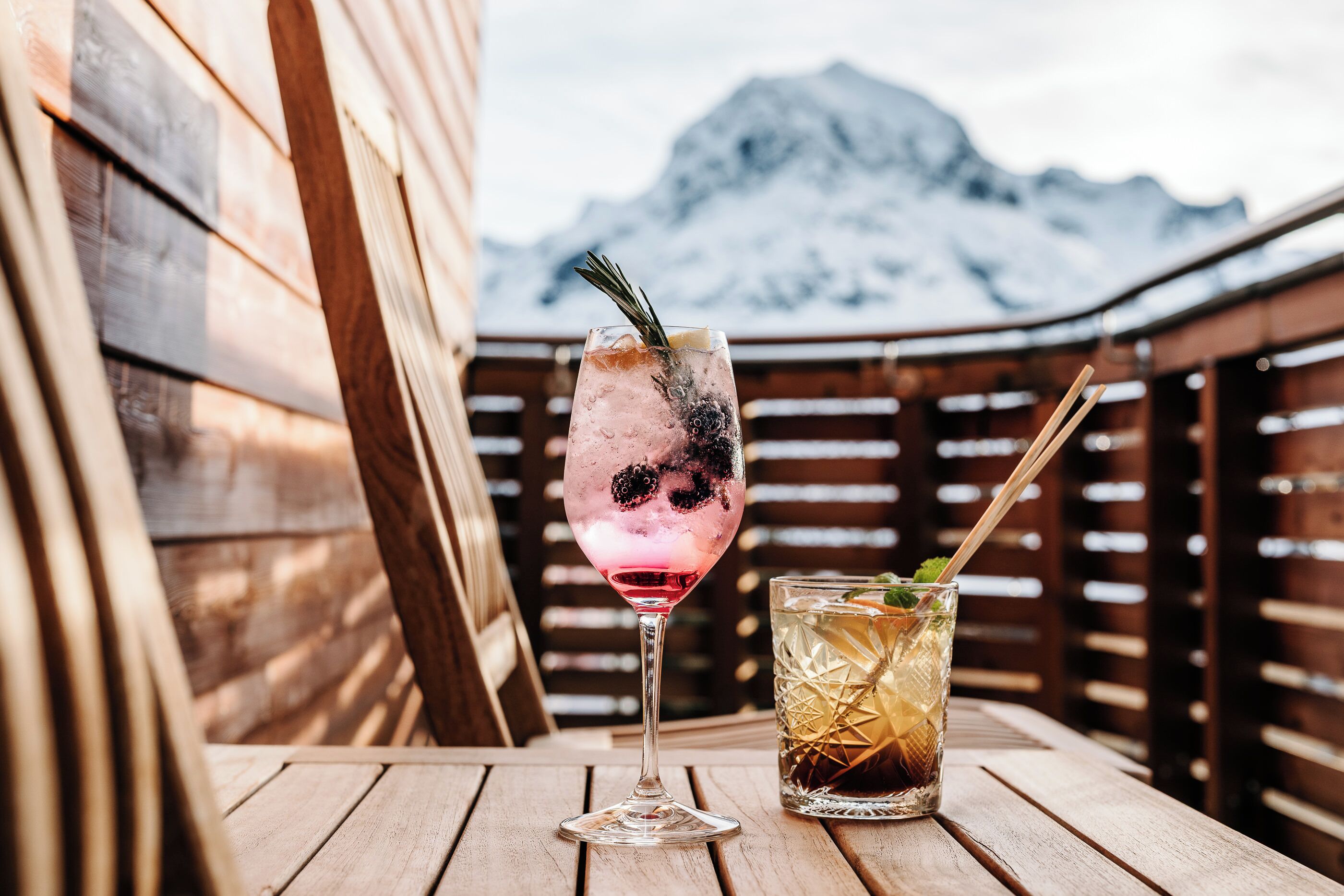 A wooden table with two drinks, one in a glass and one in a tumbler, with a mountain in the background.