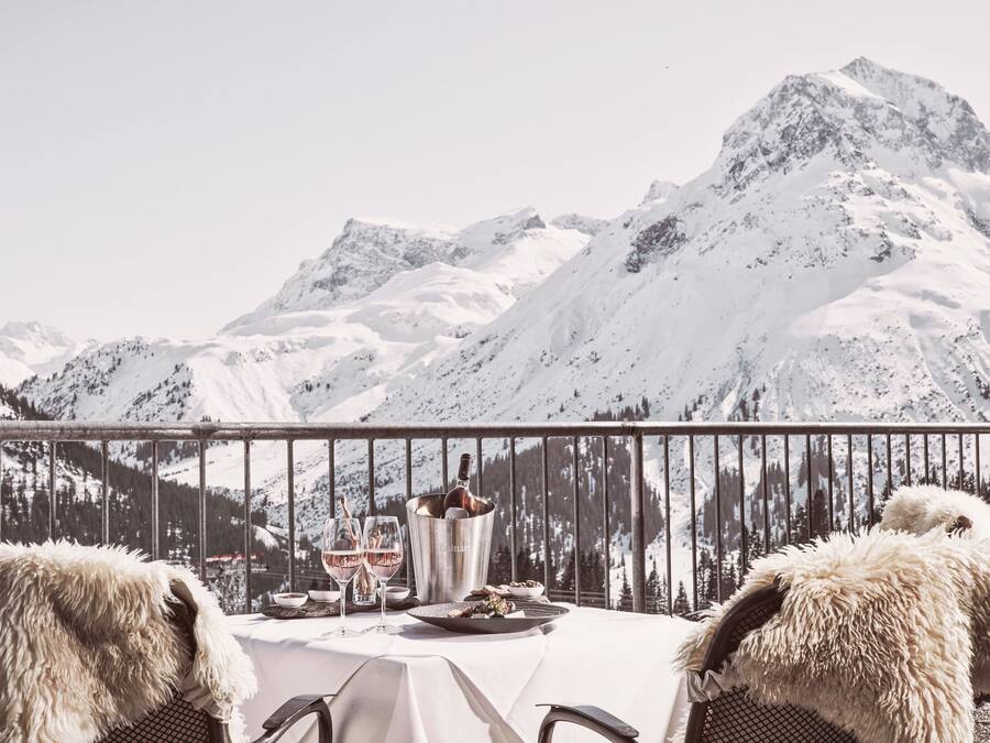 A table set for two with a bottle of champagne, glasses, and a plate of food, overlooking a snowy mountain range.