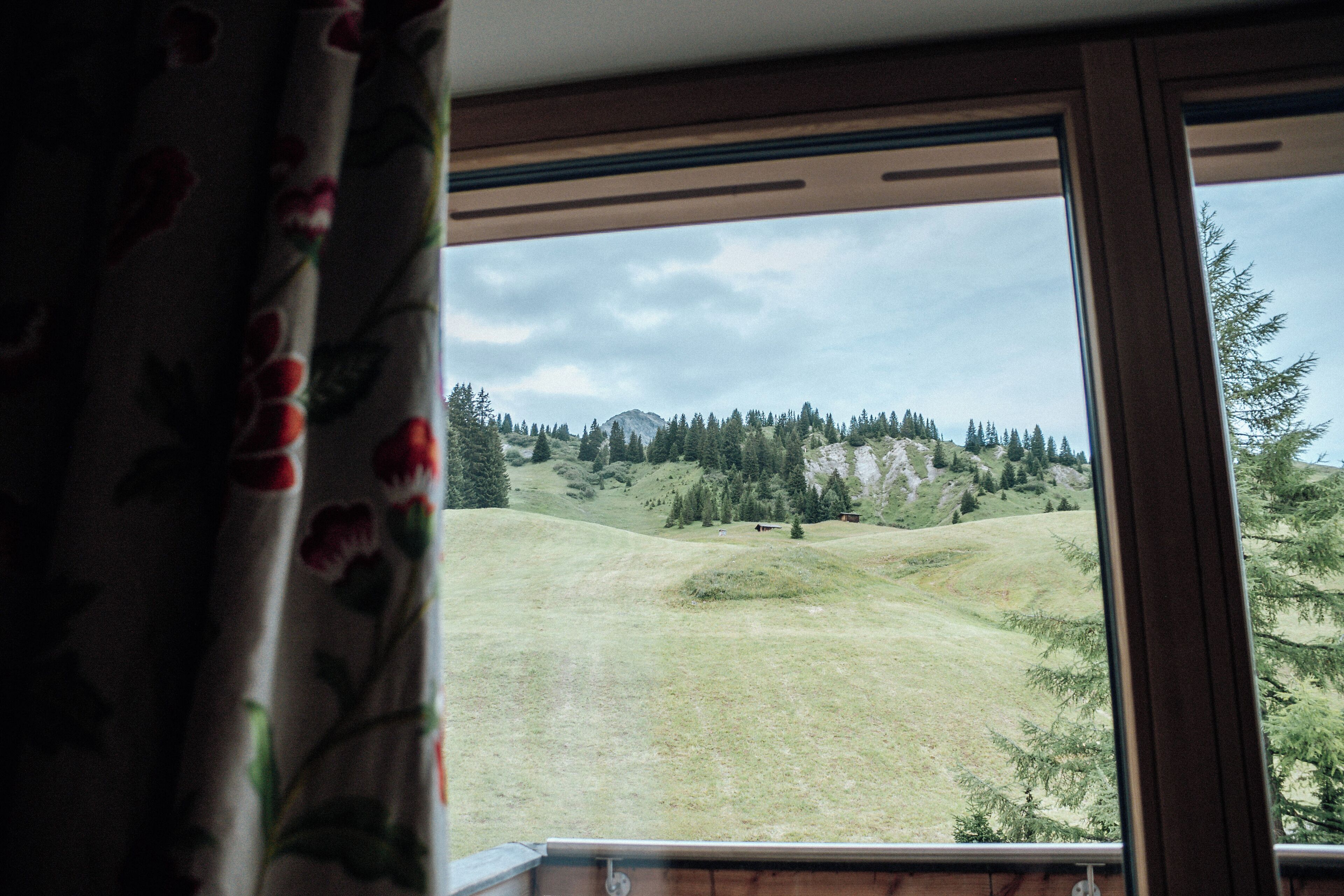 A view from a window with a curtain, overlooking a grassy landscape with trees and a mountain in the distance.