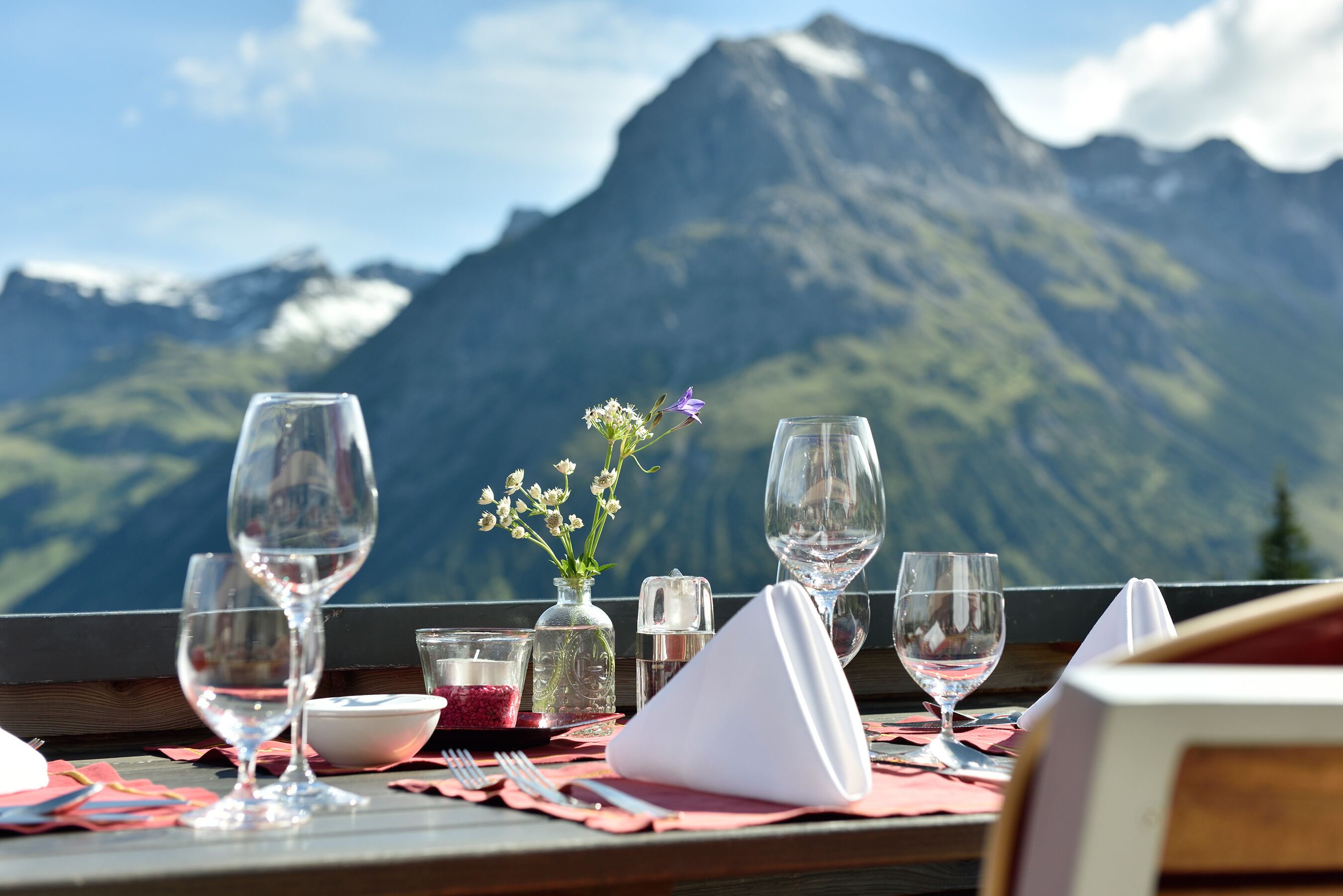 Una mesa servida con copas de vino, un florero con flores y condimentos, con vistas a una cadena montañosa.