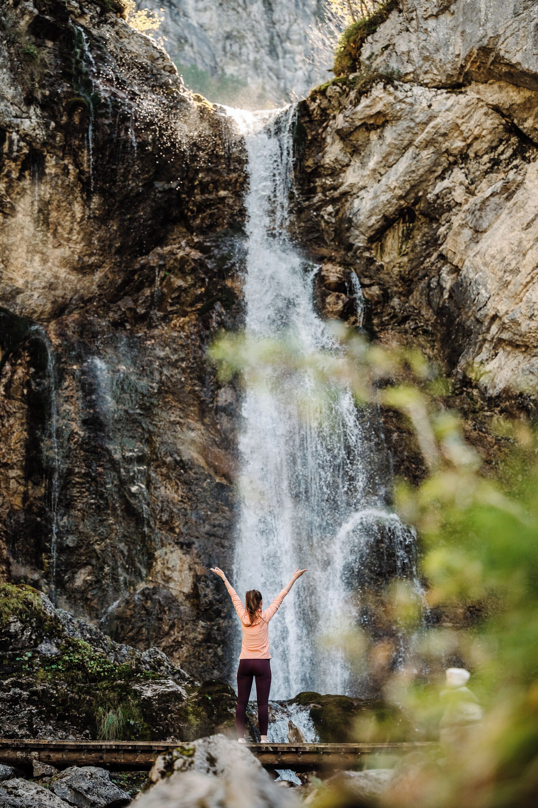A person standing with arms raised in front of a waterfall.
