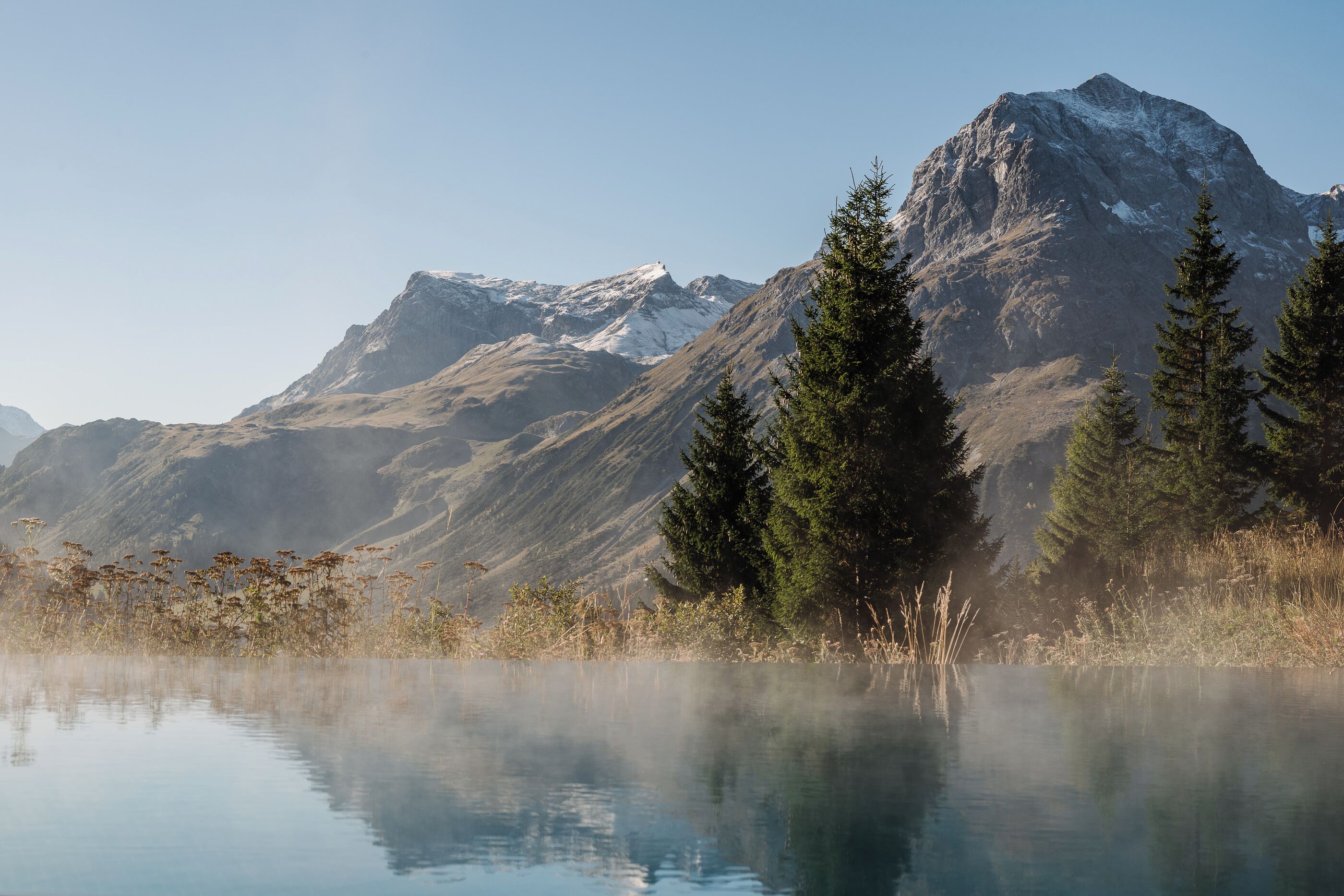 Un paisaje montañoso con un lago tranquilo que refleja las montañas, rodeado de árboles de hoja perenne.