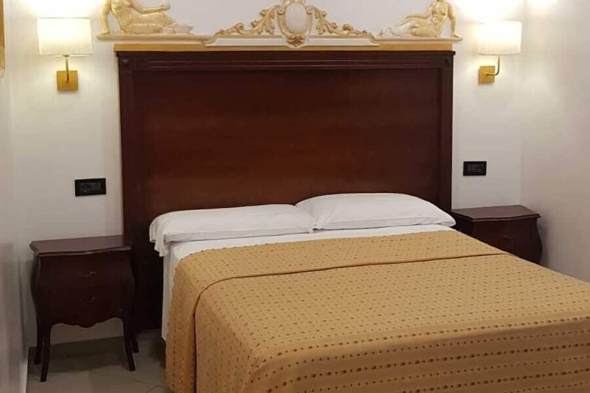 A hotel room with a wooden headboard, two bedside lamps, a bed with a beige bedspread, and a tiled floor.