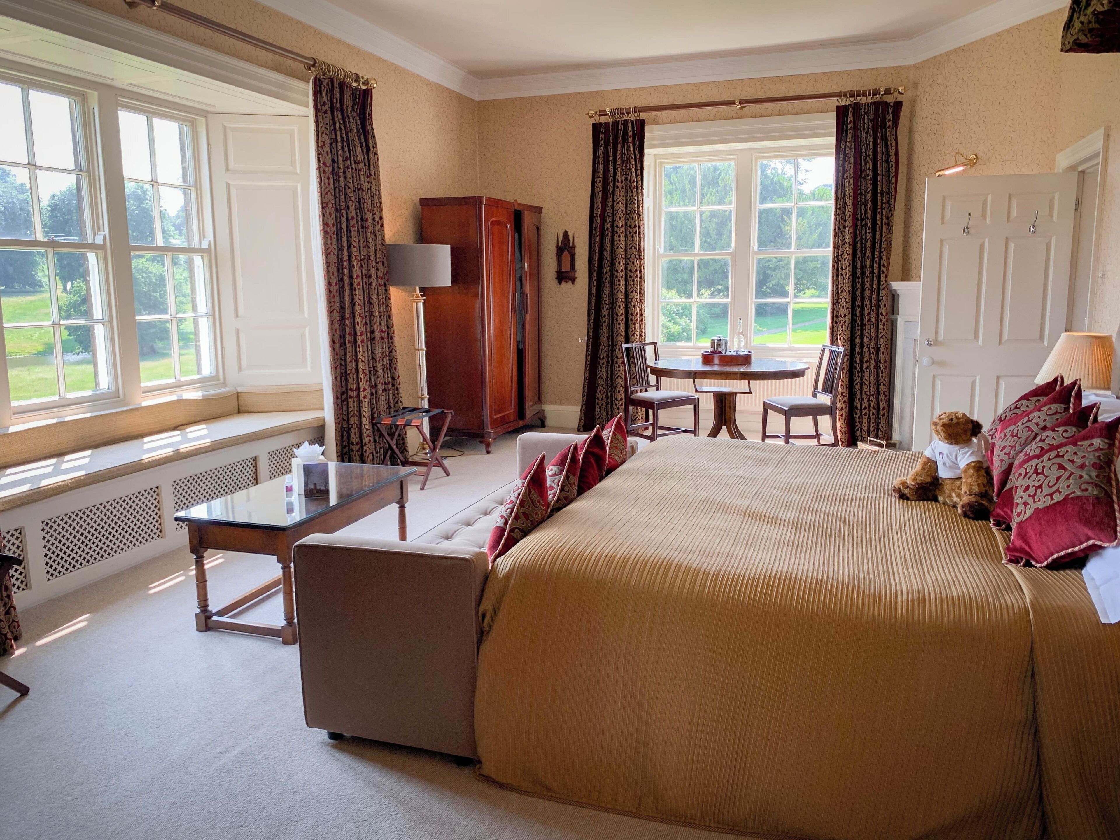 A spacious bedroom with a large bed, a seating area, a dining table, and a view of the outdoors through large windows.