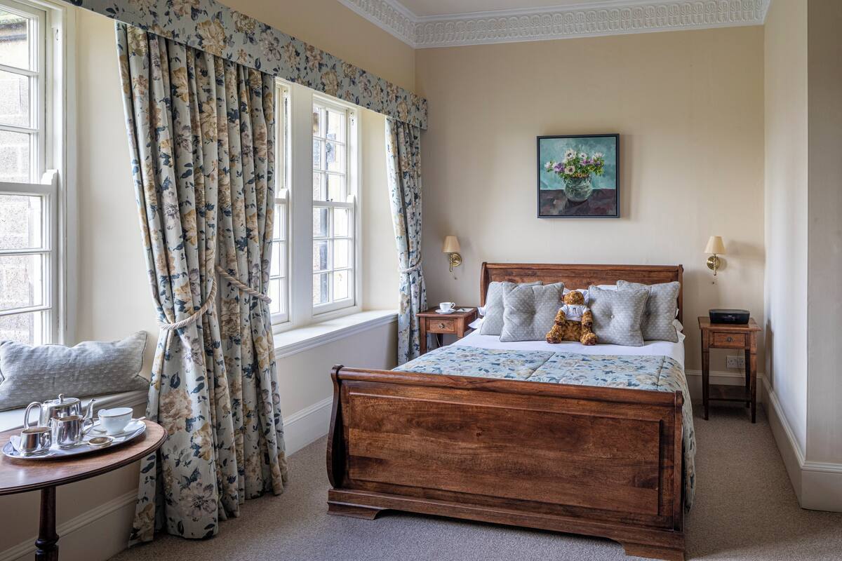 A bedroom with a wooden bed, floral curtains, a bedside table with a lamp, and a window with a view.