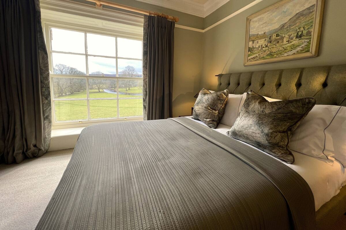 A bedroom with a large bed, a view of a green landscape through a window, and a painting on the wall.