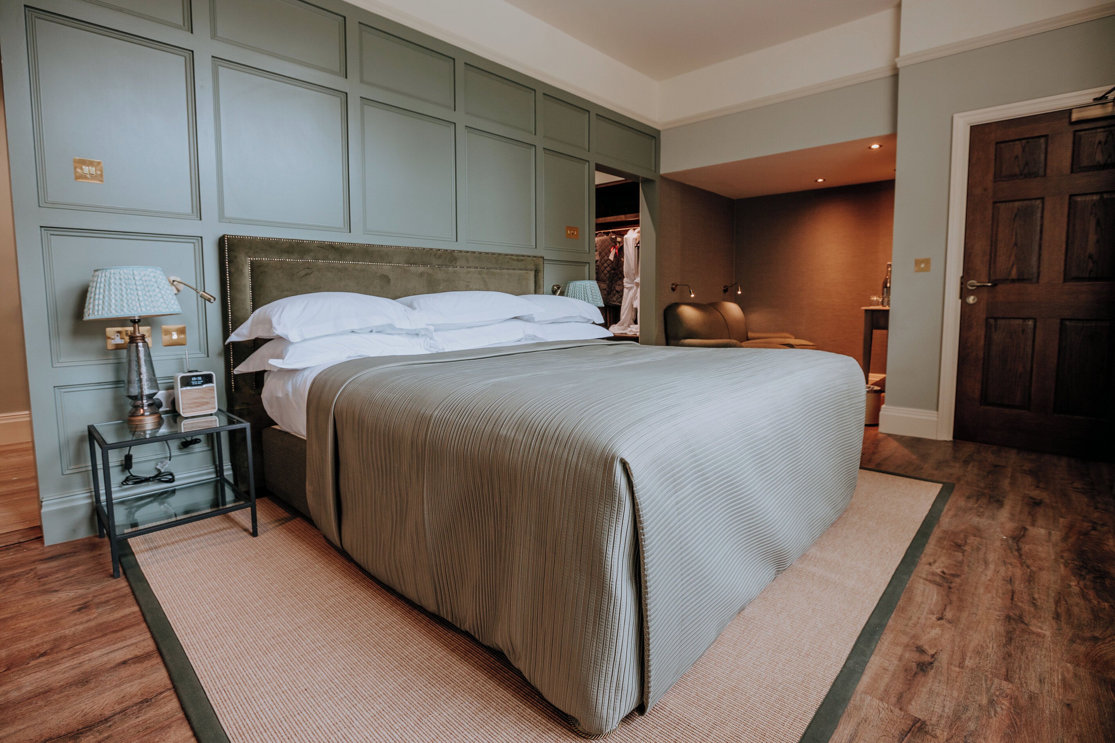 A neatly made bed with a grey headboard, a bedside table with a lamp, and a wooden floor.