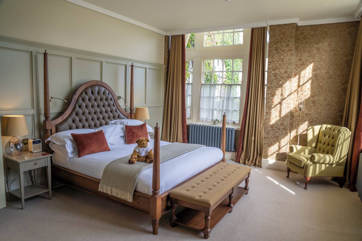 A four-poster bed with a teddy bear, a bench, a chair, and a nightstand with a lamp.
