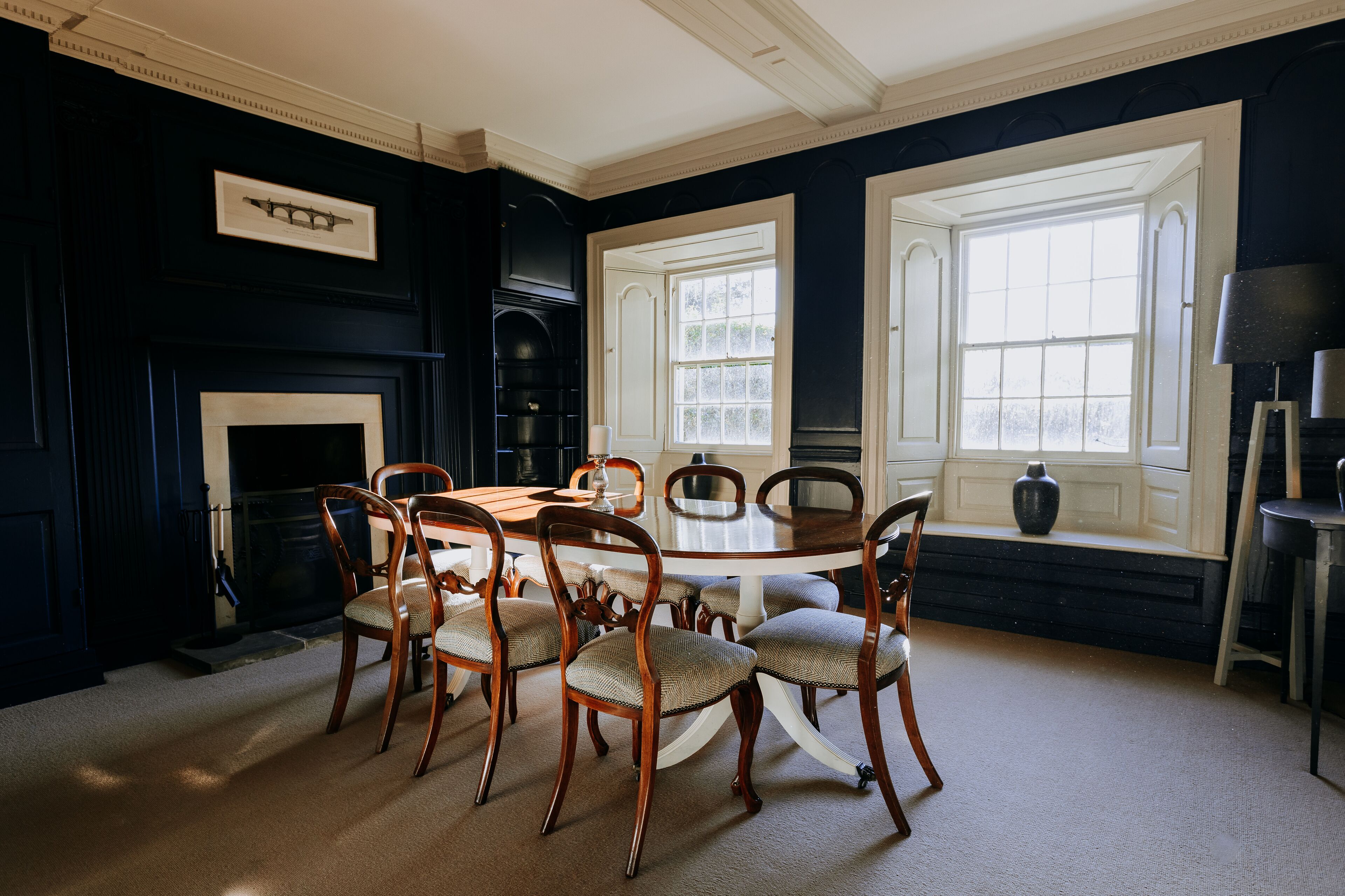A dining room with a round table and six chairs, a fireplace, and two large windows.
