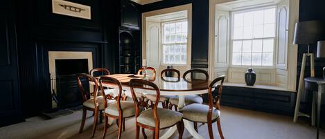 A dining room with a round table and six chairs, a fireplace, and two large windows.