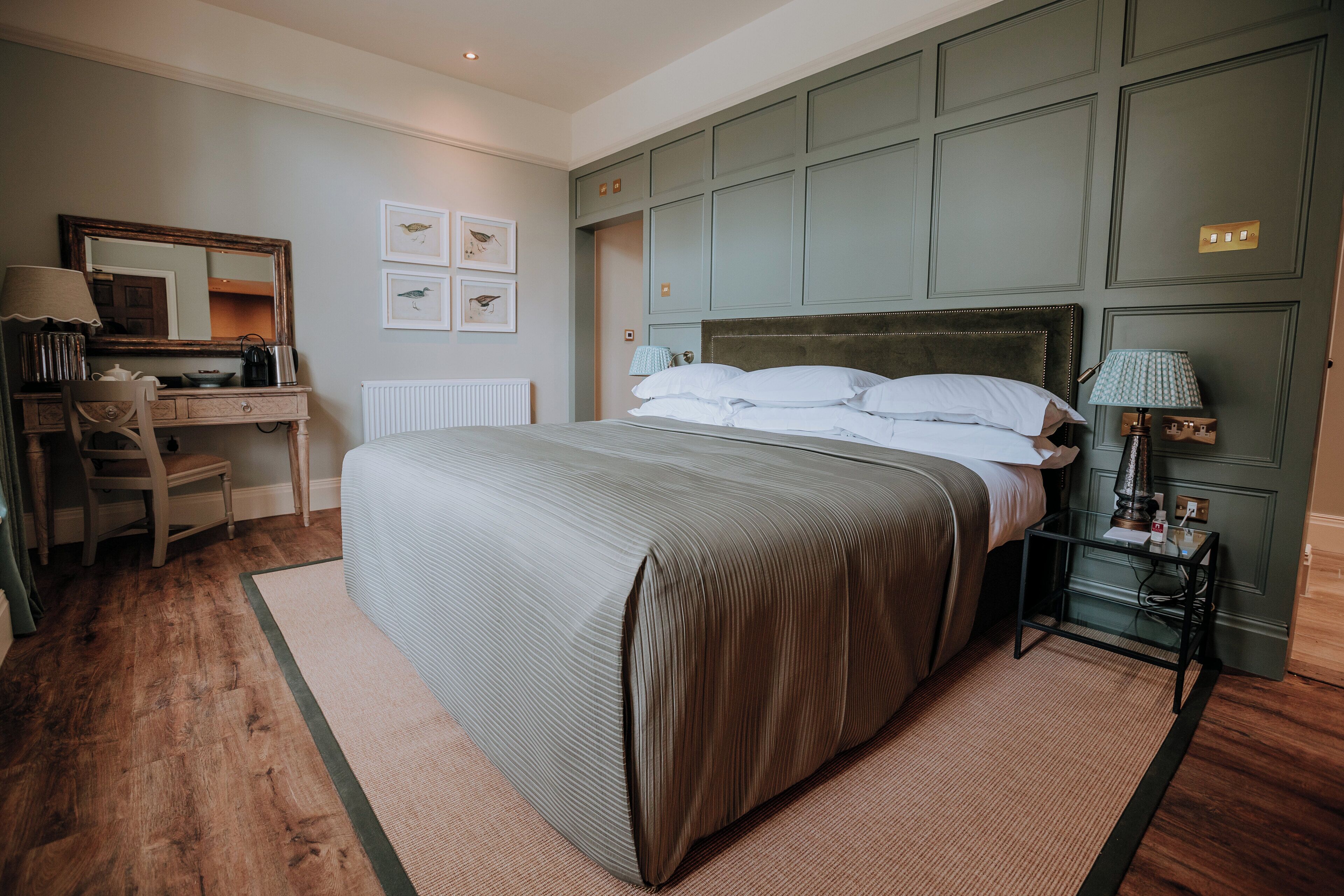 A bedroom with a large bed, a desk with a chair, a dresser, and a nightstand with a lamp.