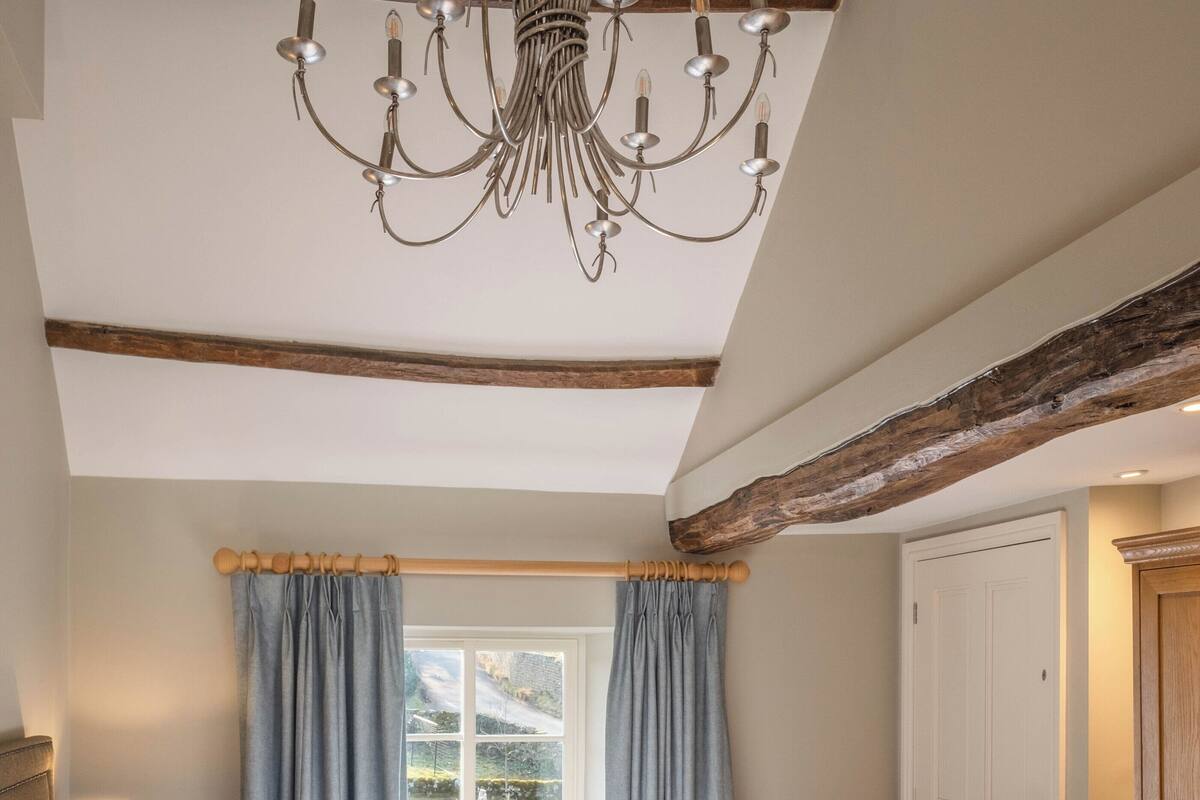 A bedroom with a large bed, a chandelier, a window with curtains, and a wooden ceiling beam.