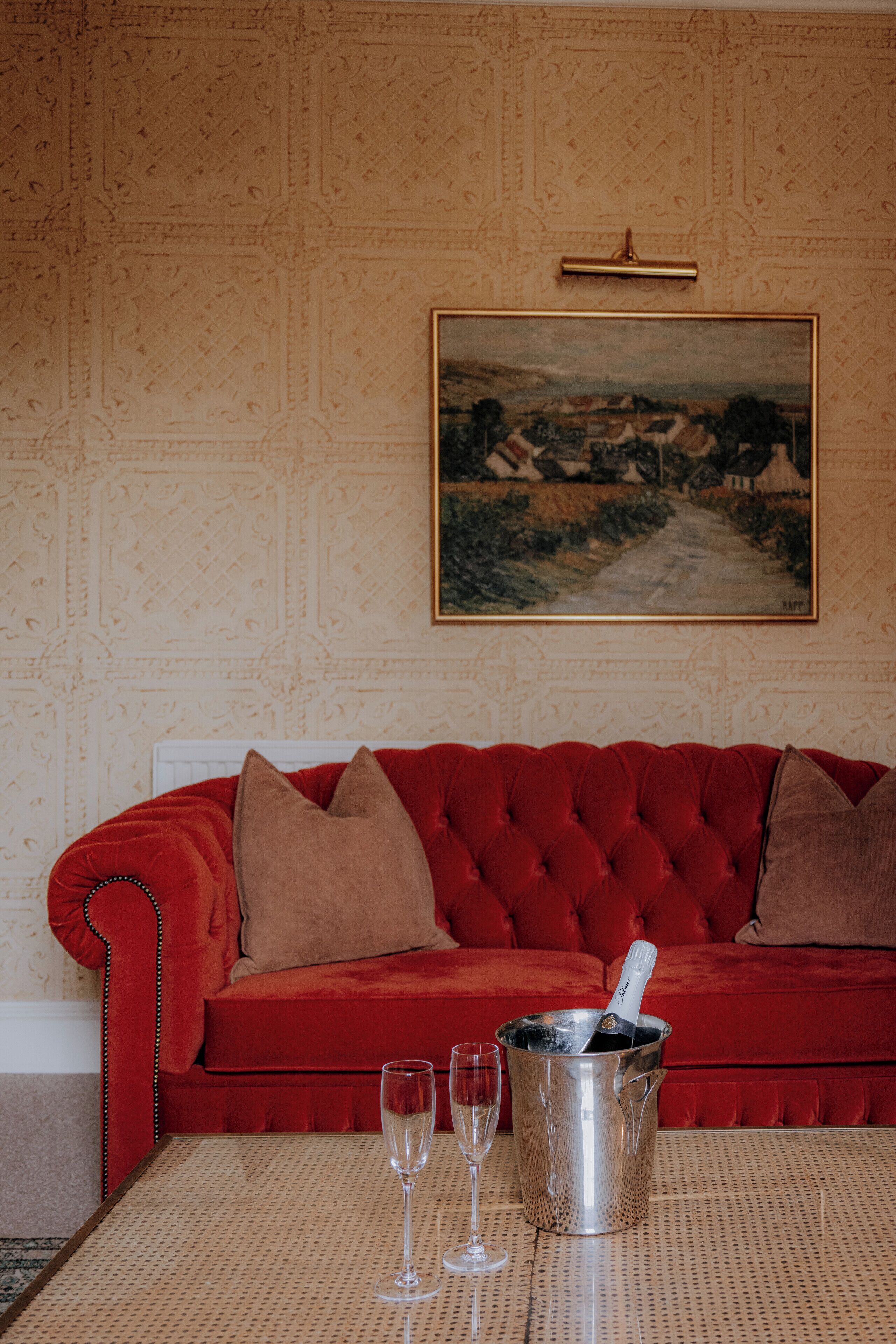 A red velvet sofa with two pillows, a champagne bucket with a bottle, and two glasses on a woven table.