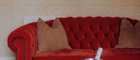 A red velvet sofa with two pillows, a champagne bucket with a bottle, and two glasses on a woven table.