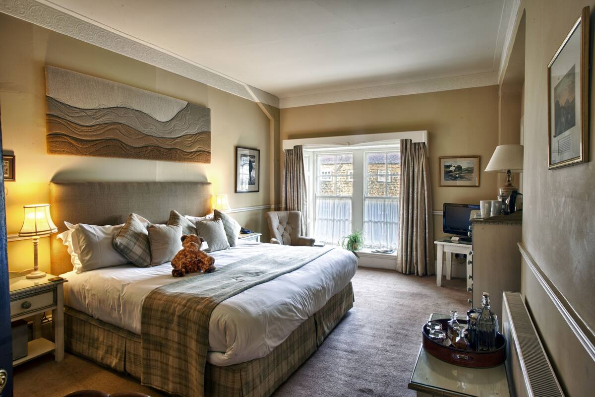 A hotel room with a large bed, a desk with a computer, a chair, a window with curtains, and a wall-mounted artwork.