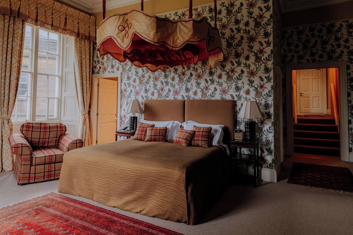 A bedroom with a large bed, a plaid armchair, a nightstand, and a window with curtains.