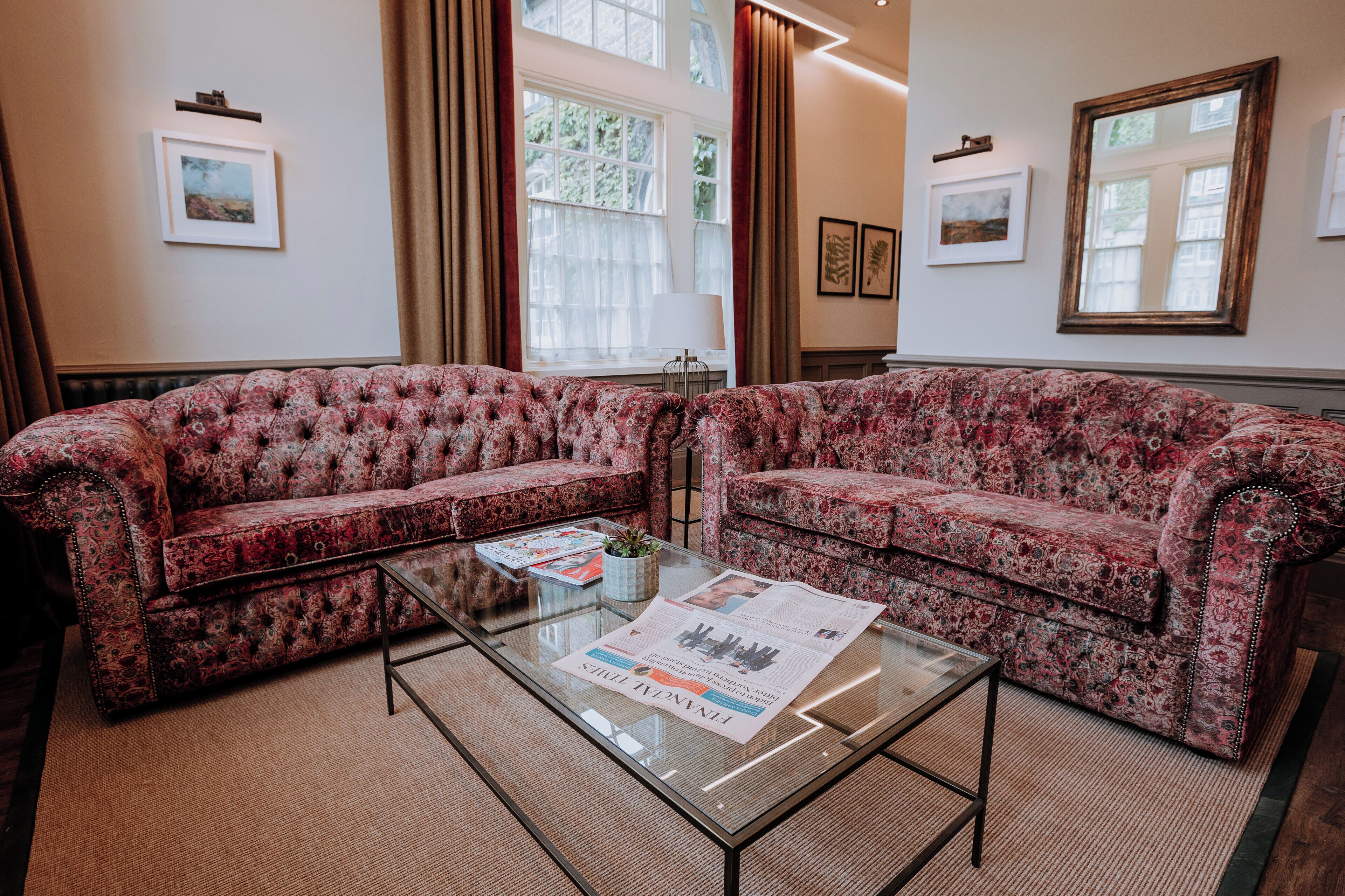 A room with two patterned sofas, a glass coffee table with a newspaper, and large windows with curtains.