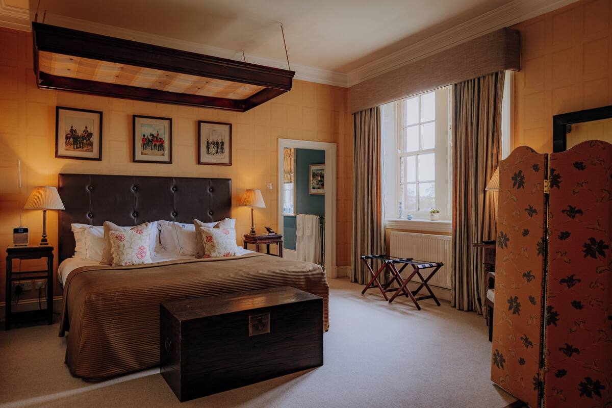 A hotel room with a large bed, two bedside tables, a nightstand, a dresser, a window with curtains, and framed pictures on the wall.