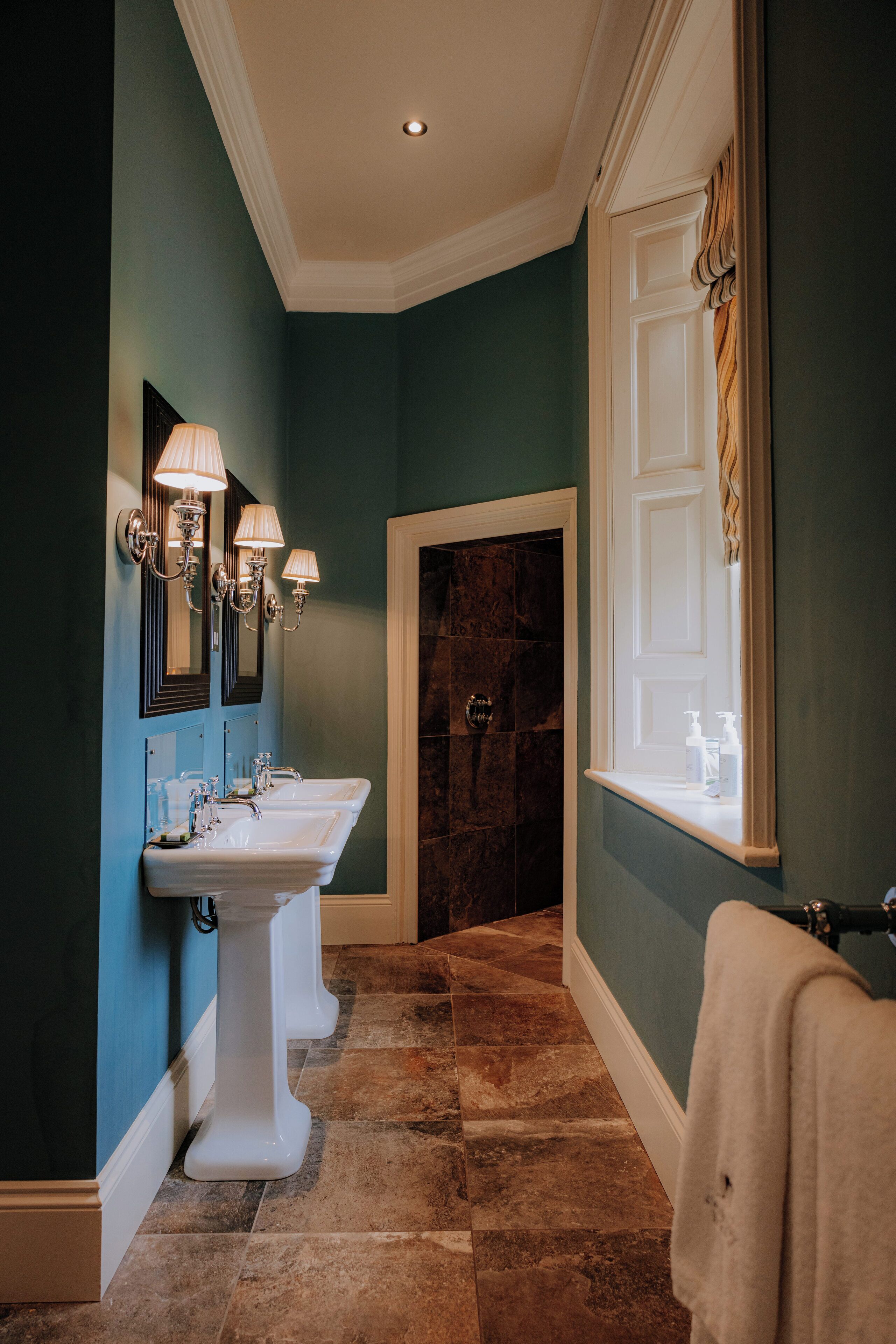 A bathroom with two sinks, wall-mounted lighting, a window with curtains, and a marble shower area.