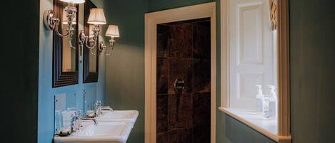 A bathroom with two sinks, wall-mounted lighting, a window with curtains, and a marble shower area.