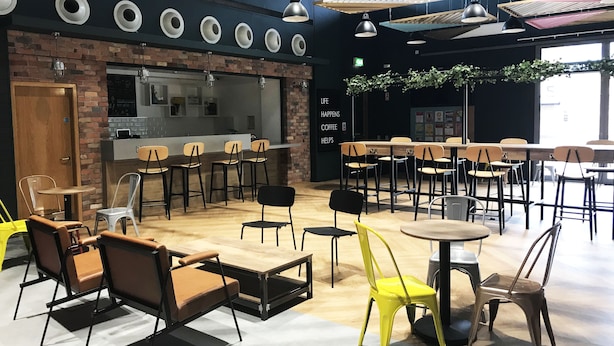 Una cafeteria moderna amb una paret de maó, taules i cadires de fusta. Hi ha una zona de bar amb tamborets i un rètol que diu "Life Happens Coffee Helps".