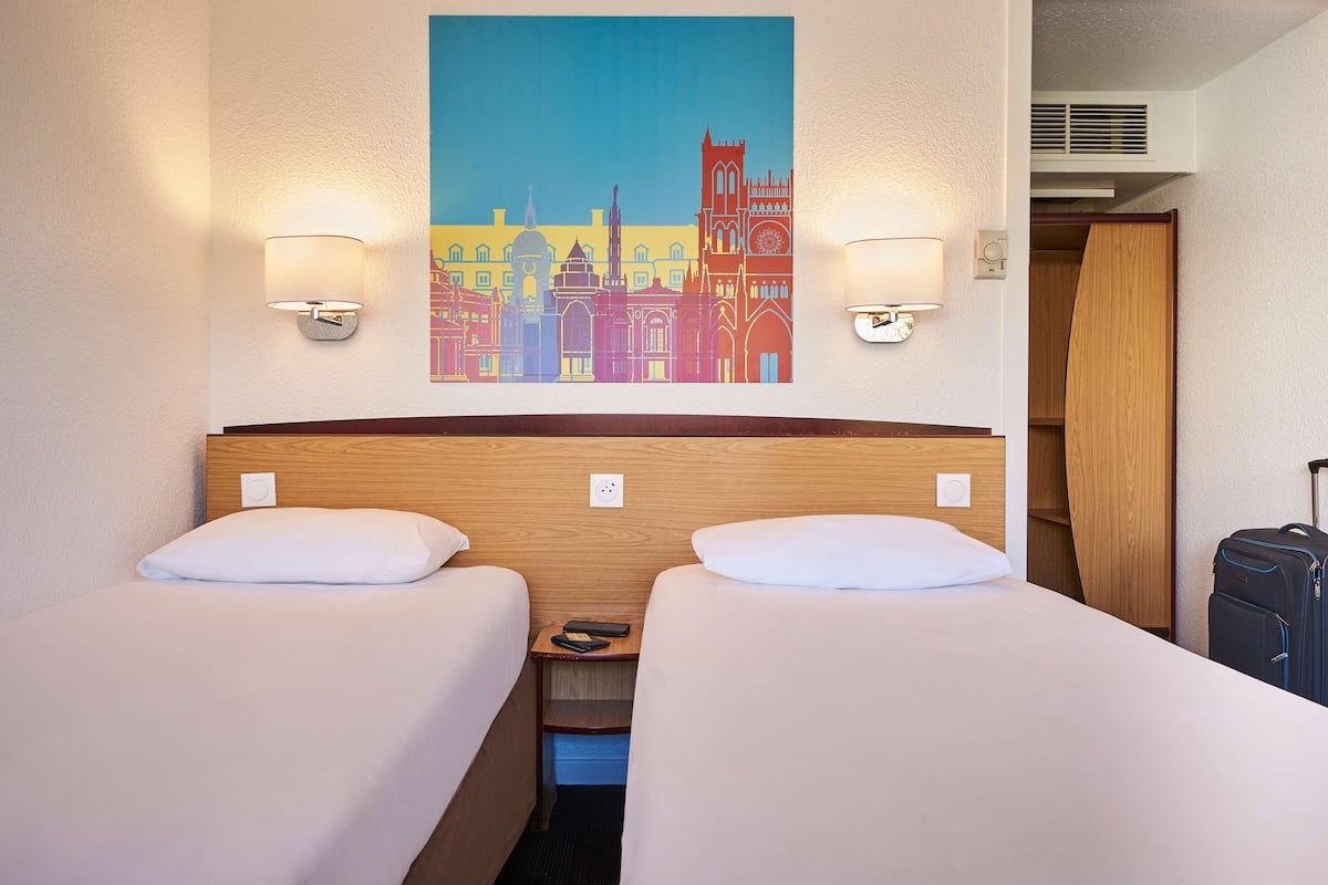 A hotel room with two beds, a painting of a cityscape, and a suitcase.