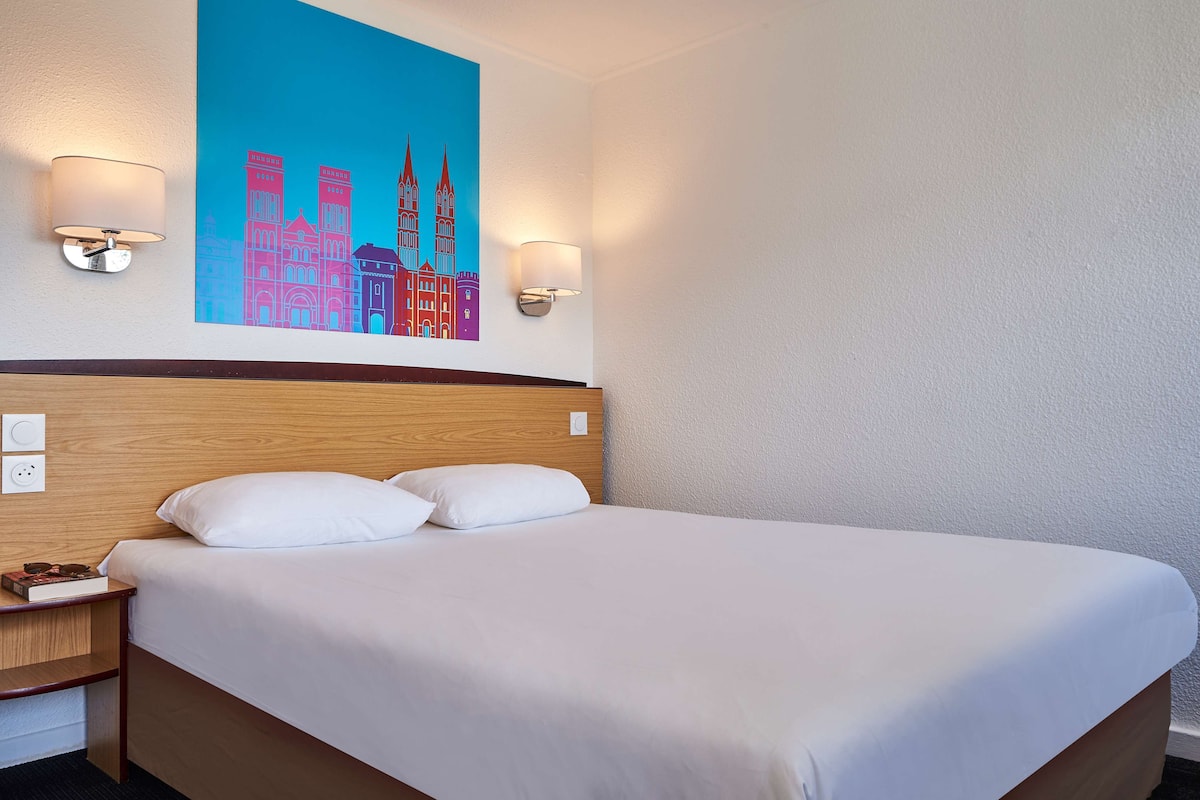 A hotel room with a large bed, two wall-mounted lamps, and a cityscape painting above the bed.