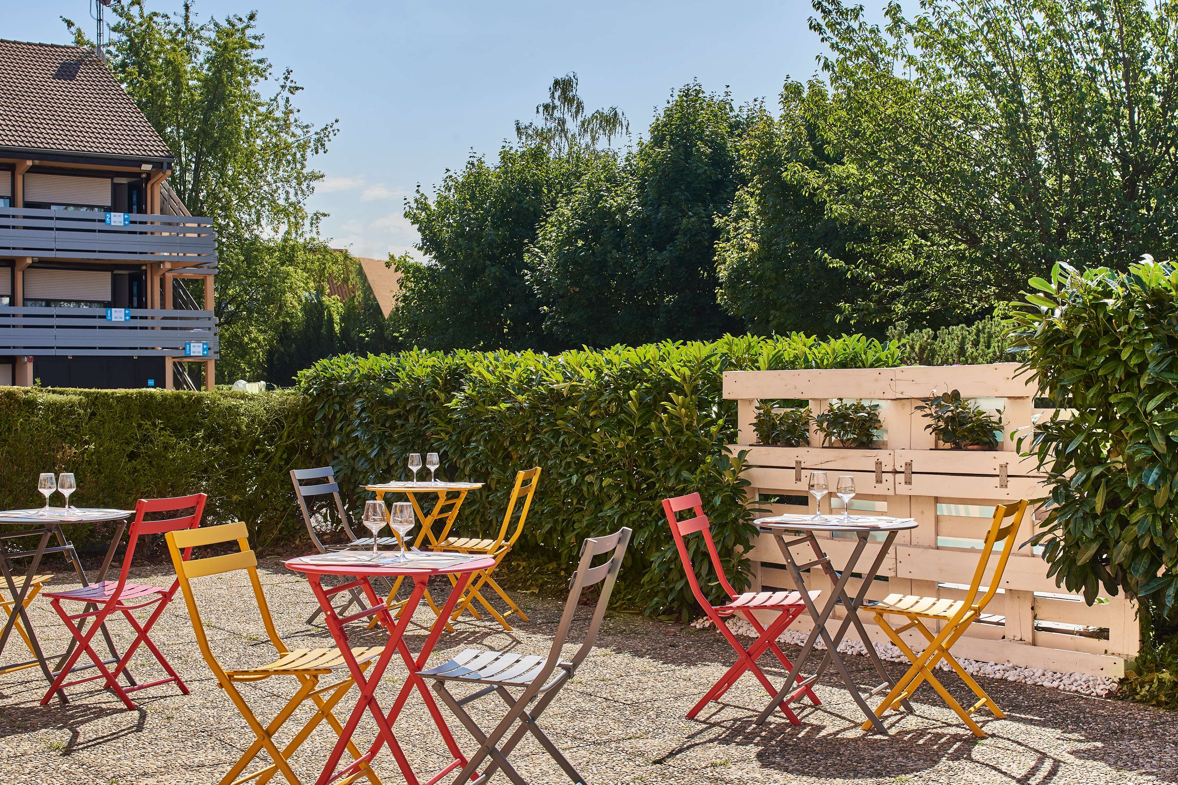 Outdoor seating with tables and chairs, a building in the background, and a hedge.