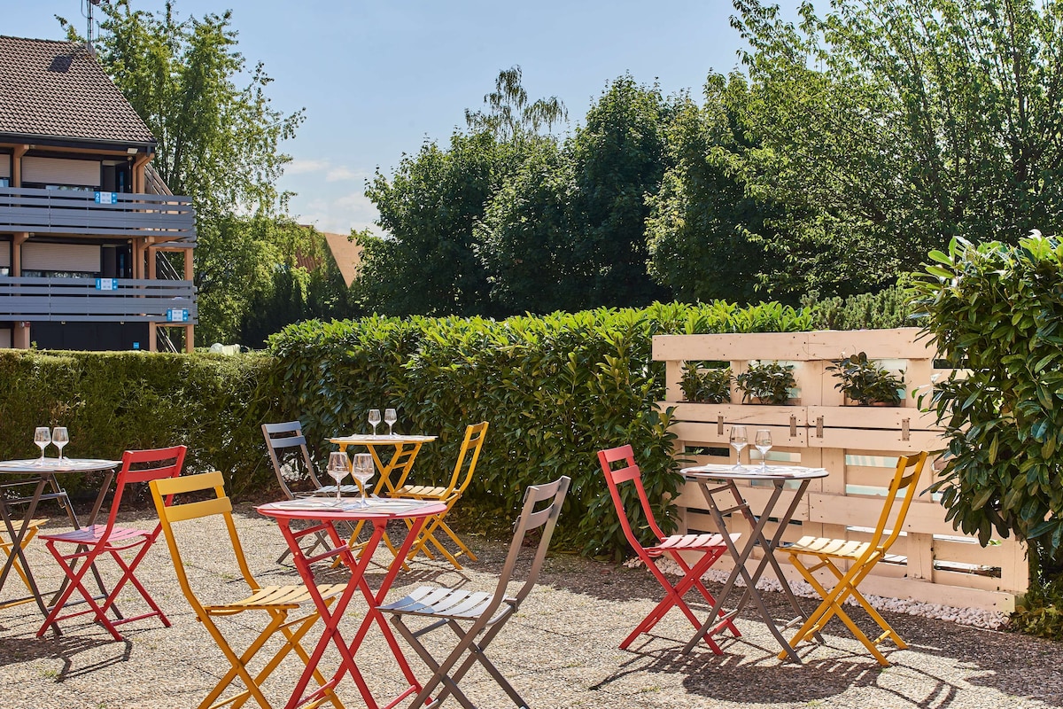 An outdoor seating area with colorful chairs and tables, surrounded by greenery and a building in the background.