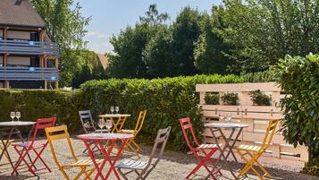 Outdoor seating with tables and chairs, a building in the background, and a hedge.