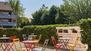 An outdoor seating area with colorful chairs and tables, surrounded by greenery and a building in the background.