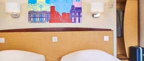 A hotel room with two beds, a wooden headboard, a wall-mounted painting of a cityscape, two wall sconces, a bedside table with a lamp, and a suitcase.
