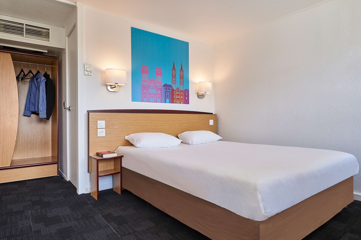 Habitación de hotel con cama, mesita de noche, cabecera de madera, armario con ropa colgada y un cuadro en la pared que representa un paisaje urbano.